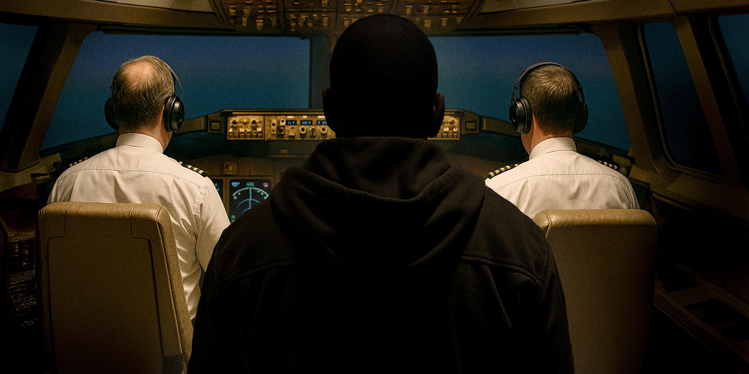 Ein schlecht beleuchteter Mann steht mit dem Rücken zur Kamera in einem Cockpit, in dem zwei Piloten sitzen